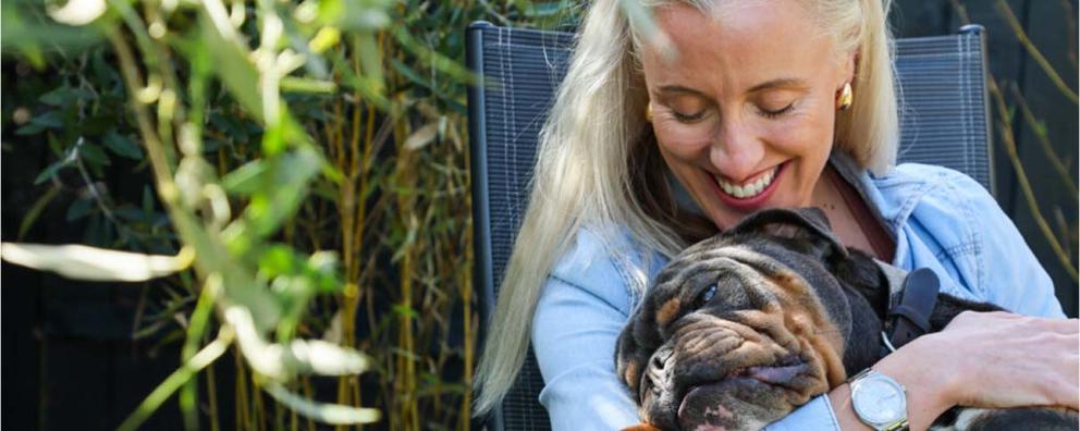 A lady with blonde hair and a blue shirt, smiling down at Bernie, a black and brown bulldog breed dog, she is holding in her arms.